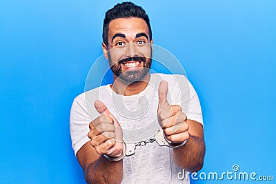 Young hispanic man wearing prisoner handcuffs smiling happy and positive, thumb up doing excellent and approval sign Stock Photo