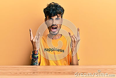 Young hispanic man wearing tshirt with happiness word message sitting on the table shouting with crazy expression doing rock Stock Photo