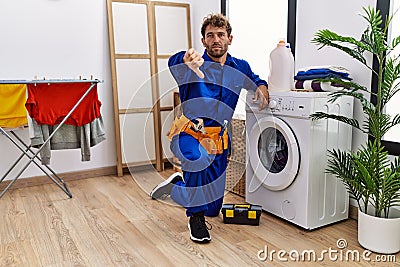 Young hispanic man working on washing machine with angry face, negative sign showing dislike with thumbs down, rejection concept Stock Photo