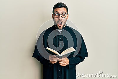 Young hispanic priest man holding bible afraid and shocked with surprise and amazed expression, fear and excited face Stock Photo