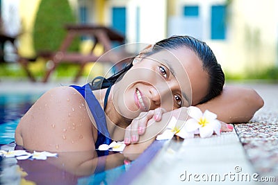 Young hispanic woman in blue dress relaxing by the swimming pool surrounded by flowers Stock Photo