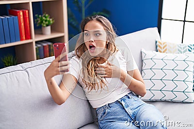 Young hispanic woman doing video call with smartphone scared and amazed with open mouth for surprise, disbelief face Stock Photo