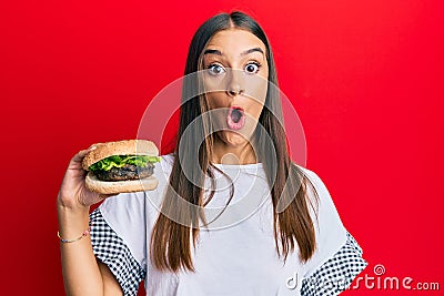 Young hispanic woman eating hamburger scared and amazed with open mouth for surprise, disbelief face Stock Photo