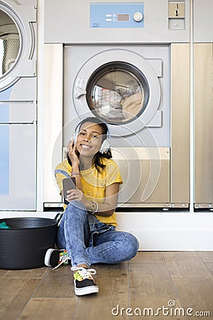 Young hispanic woman enjoying music waiting for the clothes to be washed sitting on the floor at the self-service laundry Stock Photo