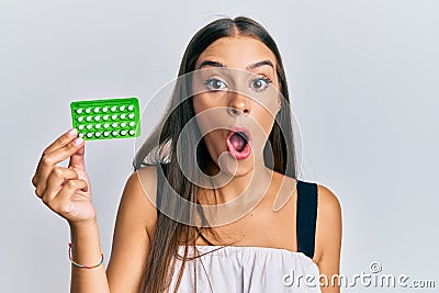 Young hispanic woman holding birth control pills scared and amazed with open mouth for surprise, disbelief face Stock Photo