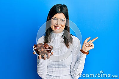 Young hispanic woman holding dates bowl smiling happy pointing with hand and finger to the side Stock Photo