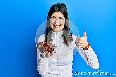 Young hispanic woman holding dates bowl smiling happy and positive, thumb up doing excellent and approval sign Stock Photo