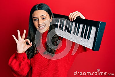 Young hispanic woman holding piano keyboard doing ok sign with fingers, smiling friendly gesturing excellent symbol Stock Photo