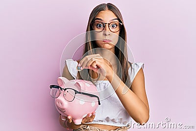 Young hispanic woman holding piggy bank with glasses and coin puffing cheeks with funny face Stock Photo