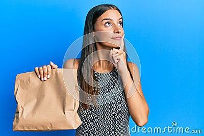 Young hispanic woman holding take away paper bag serious face thinking about question with hand on chin, thoughtful about Stock Photo