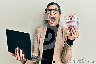 Young hispanic woman wearing business style holding laptop and swedish krone angry and mad screaming frustrated and furious, Stock Photo