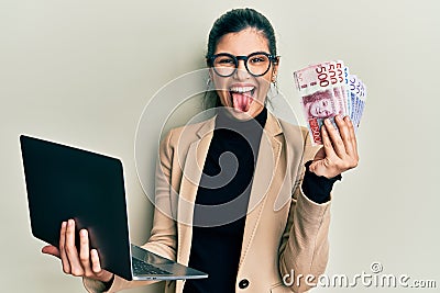 Young hispanic woman wearing business style holding laptop and swedish krone sticking tongue out happy with funny expression Stock Photo
