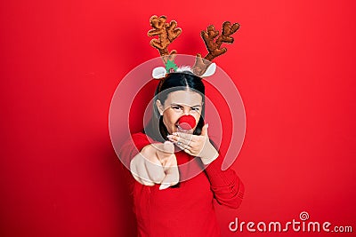Young hispanic woman wearing deer christmas hat and red nose laughing at you, pointing finger to the camera with hand over mouth, Stock Photo