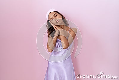 Young hispanic woman wearing sleep mask and nightgown sleeping tired dreaming and posing with hands together while smiling with Stock Photo