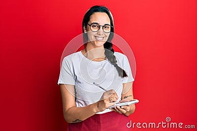 Young hispanic woman wearing waitress apron taking order smiling with a happy and cool smile on face Stock Photo