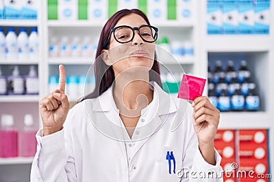 Young hispanic woman working at pharmacy holding condom smiling with an idea or question pointing finger with happy face, number Stock Photo