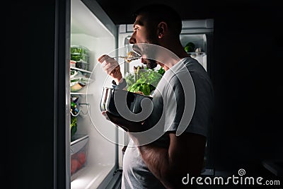 Young hungry man eating food at night from open fridge. Midnight snack and food addiction Stock Photo