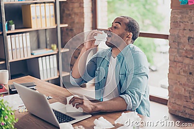 Young ill afro student is sneezing at work place in modern office, a lot of paper napkins on desktop and in his arm Stock Photo