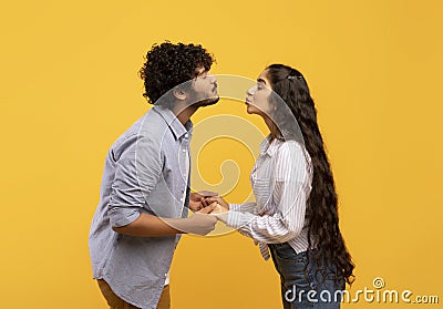 Young indian couple reaching each other with lips, ready to kiss over yellow background, side view Stock Photo