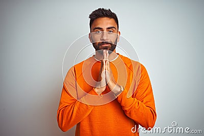 Young indian man wearing orange sweater over isolated white background praying with hands together asking for forgiveness smiling Stock Photo