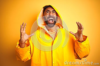 Young indian man wearing raincoat with hood standing over isolated yellow background crazy and mad shouting and yelling with Stock Photo