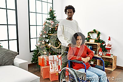 Young interracial couple with woman sitting on wheelchair by christmas tree skeptic and nervous, frowning upset because of problem Stock Photo