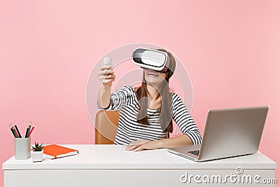 Young joyful woman in headset of virtual reality on head using remote control sit work at white desk with pc laptop Stock Photo