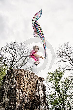 Young joyful woman is posing on the tree stump with waving scarf Stock Photo