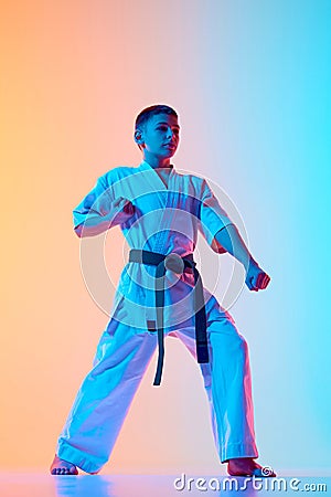 Young karateka in a defensive posture, teen boy practicing against gradient orange blue background in neon light Stock Photo
