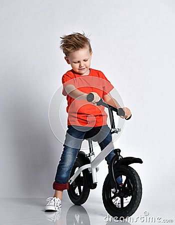 Young kid boy in orange t-shirt and blue jeans biker sits on his bicycle and looks at it like check if it is ready for race Stock Photo