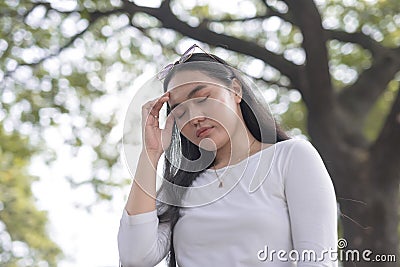 A young lady standing outside the park closes her eyes and touches the temple of her head as she experiences headache Stock Photo