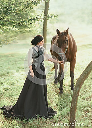 A young lady in a vintage dress strolls through the forest with her horse. The girl has a white blouse, a jabot, a tie Stock Photo