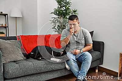 Young latin man and dog sitting on the sofa at home feeling unwell and coughing as symptom for cold or bronchitis Stock Photo