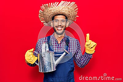 Young latin man wearing farmer hat and gloves holding watering can smiling with an idea or question pointing finger with happy Stock Photo