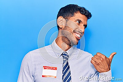 Young latin man wearing sticker with hello my name is message pointing thumb up to the side smiling happy with open mouth Stock Photo