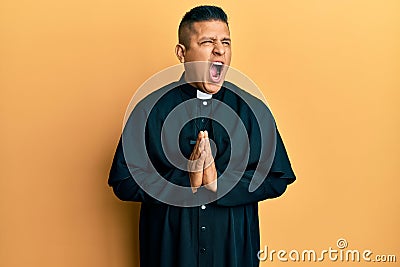 Young latin priest man praying with hands together angry and mad screaming frustrated and furious, shouting with anger Stock Photo