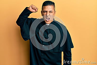 Young latin priest man standing over yellow background angry and mad raising fist frustrated and furious while shouting with anger Stock Photo