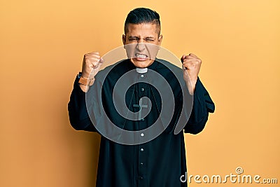 Young latin priest man standing over yellow background angry and mad raising fists frustrated and furious while shouting with Stock Photo