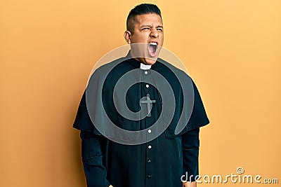 Young latin priest man standing over yellow background angry and mad screaming frustrated and furious, shouting with anger Stock Photo