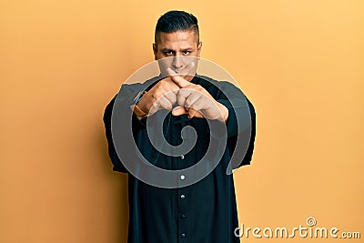 Young latin priest man standing over yellow background rejection expression crossing fingers doing negative sign Stock Photo