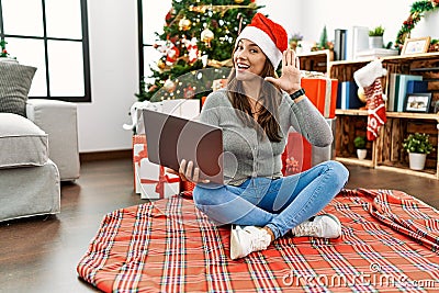 Young latin woman using laptop sitting by christmas tree waiving saying hello happy and smiling, friendly welcome gesture Stock Photo