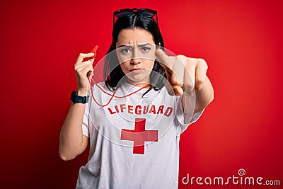 Young lifeguard woman wearing guard equipement holding whistle over red background pointing with finger to the camera and to you, Stock Photo