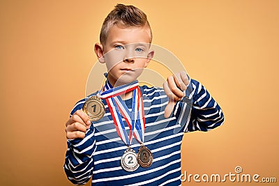 Young little caucasian winner kid wearing award competition medals over yellow background with angry face, negative sign showing Stock Photo