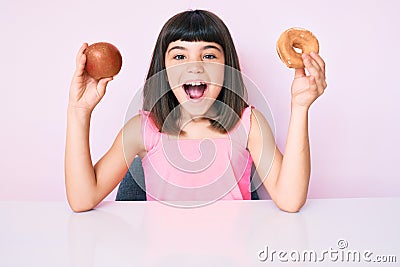 Young little girl with bang holding red apple and donut sitting on the table celebrating crazy and amazed for success with open Stock Photo