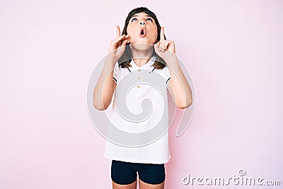 Young little girl with bang wearing sportswear amazed and surprised looking up and pointing with fingers and raised arms Stock Photo
