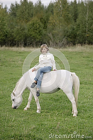 Young little girl in a white sweater and jeans sitting cross-legged on a white horse. Lifestyle portrait Stock Photo