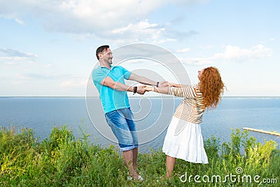 Couple dancing in grass over lake and sky background. Shot of attractive young red hair woman holding hands of the bearded man. Stock Photo