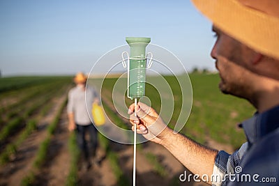 Young male agronomist holding up rain gauge with farmer using hand sprayer in background Stock Photo