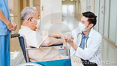 Young male Asian doctor crouch down holding hand and talk to the senior adult patient on wheelchair in hospital hallway Stock Photo