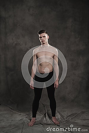 A young male ballet dancer with black leggings and a naked torso performs dance moves against a gray grunge background Stock Photo